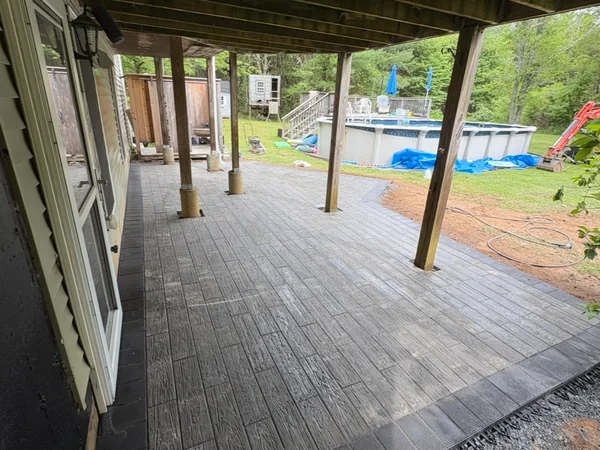 Under-Deck Paver Patio — Kingston, MA Wood-grain stamped paver patio with dark border installed under a deck beside a pool in Kingston, MA